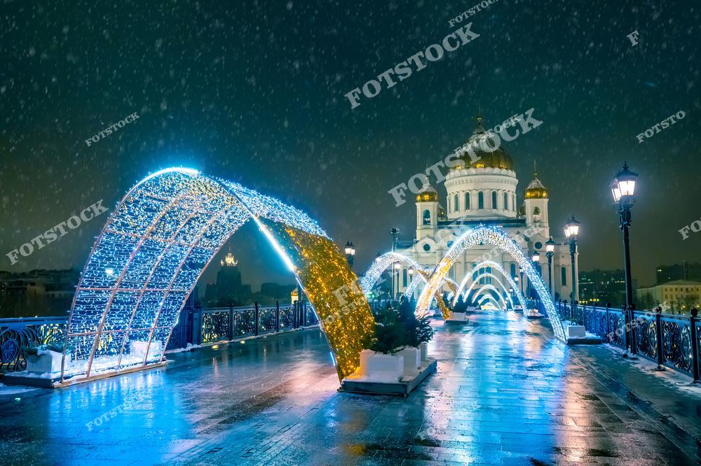 На фотографии изображён Храм Христа Спасителя, видимый с пешеходного моста, украшенного световыми арками. Падающий снег и отражения на мокрой мостовой создают атмосферу зимней сказки. Арки из гирлянд подсвечиваются синим и золотым светом, подчёркивая праздничное оформление. Вдалеке на фоне снежного неба едва заметны контуры других зданий Москвы. Это место соединяет историческую архитектуру и новогоднюю атмосферу в сердце столицы.